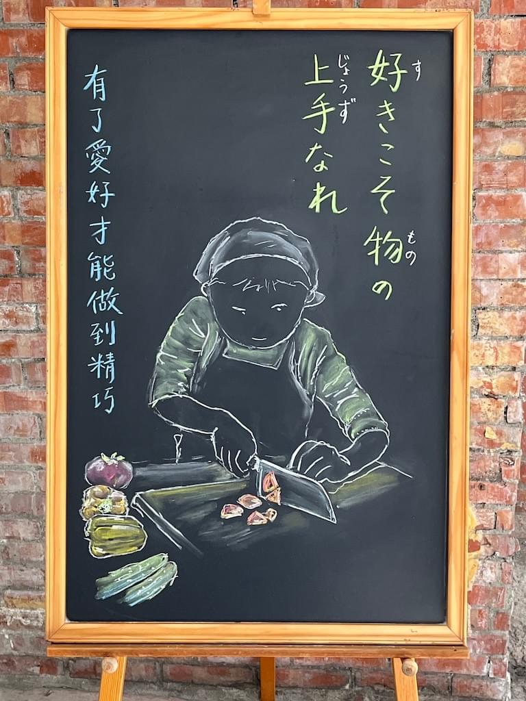 小さい黒板にチョークで絵が描いてあります。日本語のことわざ・慣用表現を紹介するための絵です。右上部に縦書き２行で「好きこそ物の／上手なれ」と書いてあります。中央に三角頭巾とエプロンをして、まな板の上でにんじんを切っている男の子のイラストが描いてあります。男の子は真剣な目で包丁を見つめています。まな板の横にきゅうり、トマト、パプリカなどの野菜が置いてあります。