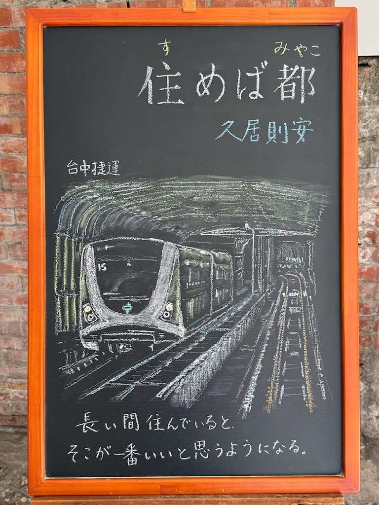 小さい黒板にチョークで絵が描いてあります。日本語のことわざを説明するための黒板です 縦長の黒板で、上部に横書き一行で「住めば都」と書いてあります。その下に中文での説明「久居則安」。その下に、台中のMRT車両のイラストが描いてあります。レールの上をこちらに向かって来る白と緑の車両で、先頭にライトが二つついています。その下に日本語で「そこに長く住んでいると、そこが一番いいと思うようになる」と書いてあります。