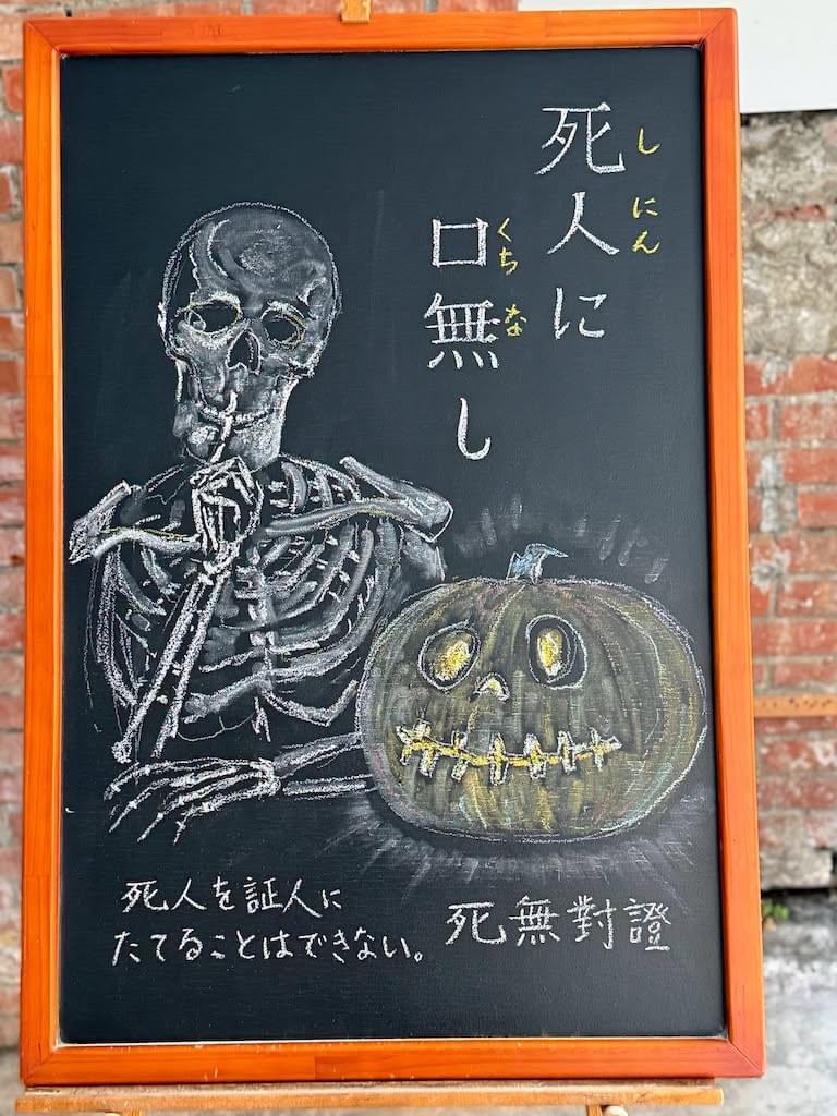 小さい黒板にチョークで絵が描いてあります。日本語のことわざを説明するための黒板です 縦長の黒板で、右上に縦書き二行で「死人に／口無し」と書いてあります。その下にかぼちゃの中をくり抜いて、お化けの顔のランタンにしたもの「ジャック・オー・ランタン」のイラスト、その右に骸骨の上半身が右手の人差し指の骨を口元にあてて「しー」というジェスチャーをしているイラストが描いてあります