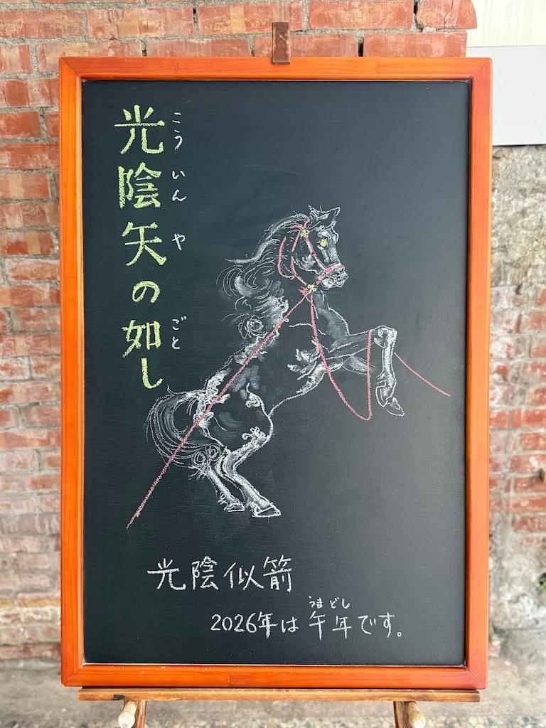 小さい黒板にチョークで絵が描いてあります。日本語のことわざを説明するための黒板です。 縦長の黒板で左上部に縦書き一行で「光陰矢の如し」と書いてあります。中央に馬のイラストが描いてあります。たてがみ、尻尾の毛をなびかせて、馬が前足を持ち上げて立ち上がっている瞬間の様子です。馬の頭部には赤っぽい頭絡がとりつけられています。そこに長い手綱が繋がれていて、左に長く延びています。右側に延びる手綱は馬の前足の上にかかっています。