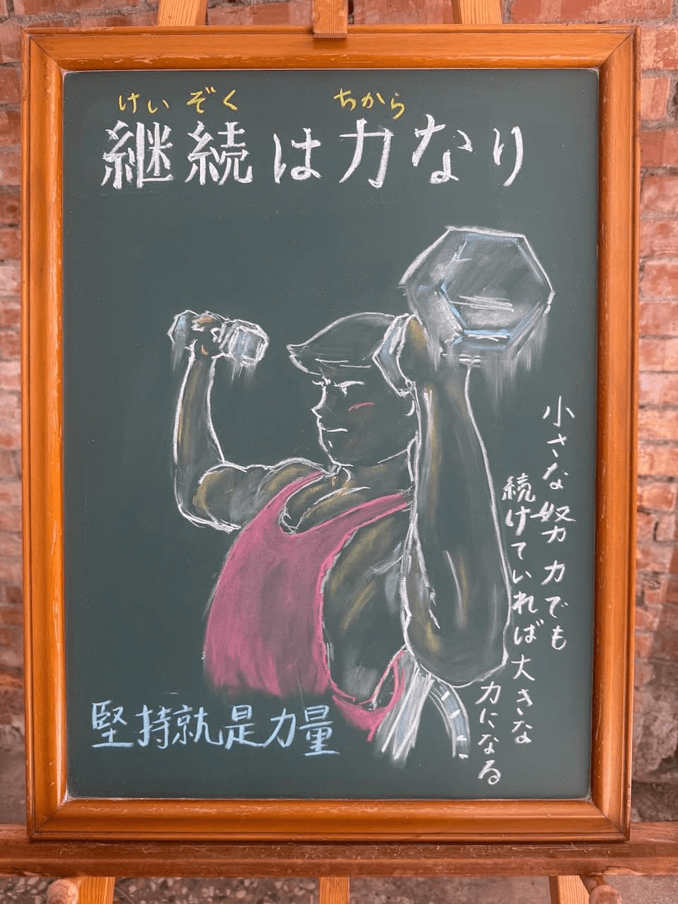 小さい黒板にチョークで絵が描いてあります。日本語のことわざを説明するための黒板です 縦長の黒板で、上部に横書き一行で「継続は力なり」と書いてあります。その下に、赤のタンクトップを着て筋トレをしている男性のイラストが描いてあります。男性は左右の手にダンベルを持ち、腕を広げた状態で頭の横の高さにあるダンベルを上に上げようと歯を食いしばっている瞬間です。 下部に横書きで中国語での意味「堅持就是力量」が書いてあります。右下に縦書きで意味の説明が書いてあります「小さな努力でも続けていれば、大きな力になる」