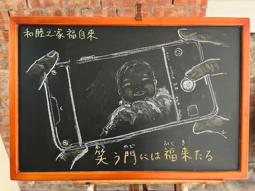 小さい黒板にチョークで絵が描いてあります。日本語のことわざ・慣用表現を紹介するための絵です。横長の枠に、スマホを両手で持って、写真を撮っている様子のイラストが大きく描いてあります。スマホの画面には笑顔の赤ちゃんが映っています。下部に「笑う門には福来たる」と書いてあり、上部に中国語で「和睦之家福自來」とあります。