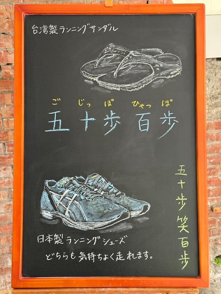 小さい黒板にチョークで絵が描いてあります。日本語のことわざ・慣用表現を紹介するための絵です。中央に横書きで「五十歩百歩」と書いてあります。その上部に、サンダルのイラストがあり、文字で「台湾製ランニングサンダル」とあります。 下部に、青いランニングシューズのイラストがあり、「日本製ランニングシューズ」と書いてあります。その下に、「どちらも気持ちよく走れます」と文字があります。