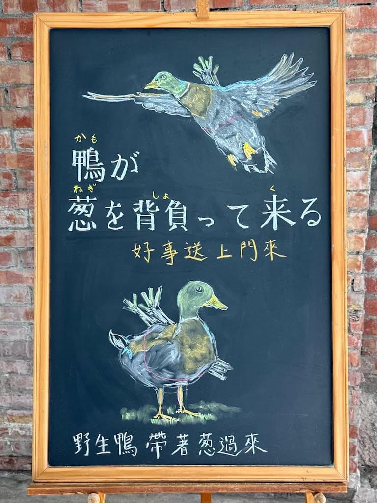 小さい黒板にチョークで絵が描いてあります。日本語のことわざを説明するための黒板です 縦長の黒板で、上部に空を飛んでいる鴨のイラストが描いてあります。背中にネギを背負っているのが見えます。 その下に横書き二行で「鴨が／葱を背負ってくる」と書いてあり、その下に横書き一行で中国語での意味「好事送上門來」が書いてあります。 下部に地面を歩く鴨のイラスト、同様に背中にネギの束を括り付けています。