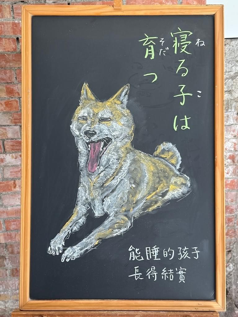 小さい黒板にチョークで絵が描いてあります。日本語のことわざを説明するための黒板です。 縦長の黒板で右上部に縦書きに行で「寝る子は／育つ」と書いてあります。その下に大きく柴犬のイラストが描いてあります。柴犬は前足を揃えて前に出して座った姿勢で、顔を持ち上げて、大きく口を開けてあくびをしている瞬間です。長い舌が見え、目はつむっています。