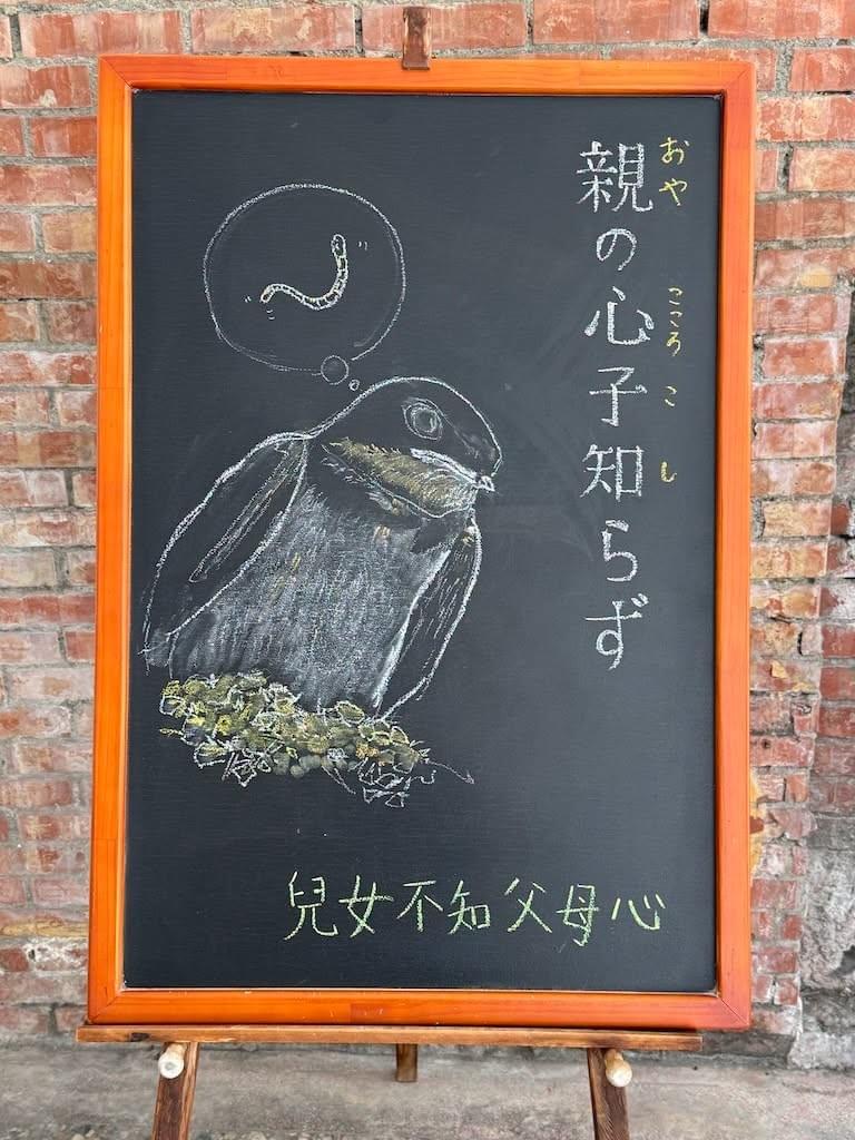 小さい黒板(縦長)にチョークで絵が描いてあります。日本語のことわざ・慣用表現を紹介するための絵です。 上部右に縦書き一行で「親の心子知らず」と書いてあります。その左に大きく燕の子のイラストが描いてあります。巣の中から体を乗り出して外を見ています。かなり成長していて体は大きくなっていますが、吹き出しの頭の中にミミズのイラストがあり、餌を待っている様子だとわかります。下部にこのことわざに相当する中文の言葉「兒女不知父母心」が書いてあります。