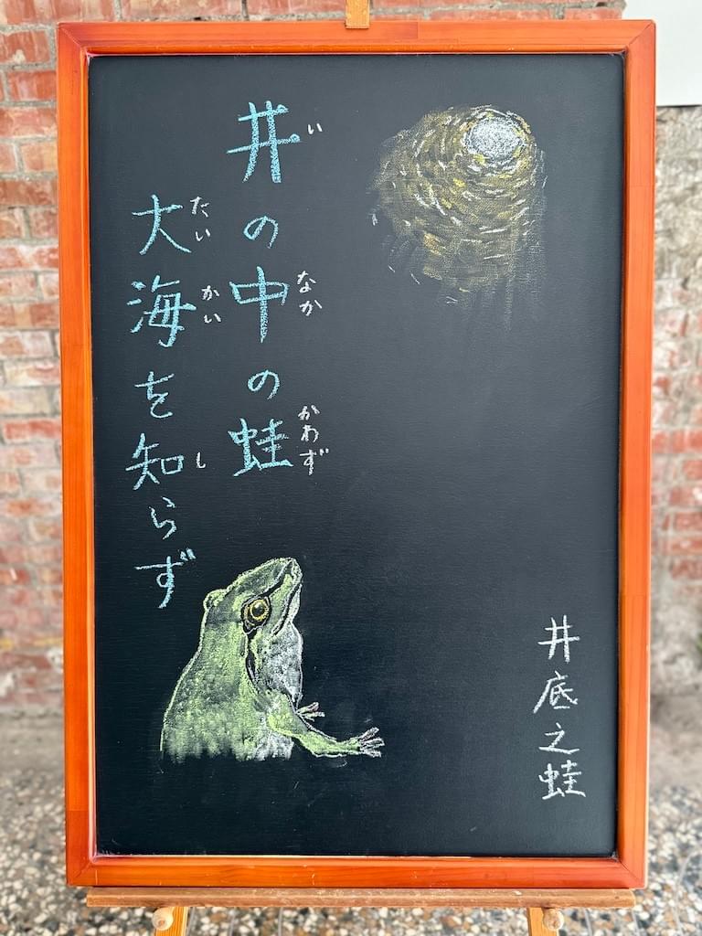 小さい黒板にチョークで絵が描いてあります。日本語のことわざ・慣用表現を紹介するための絵です。縦長の黒板で左上に縦書き二行で「井の中の蛙／大海を知らず」と書いてあります。その下に緑のカエルが上を見上げているイラストが描いてあります。カエルが見上げるのその先に井戸の入口のイラストがあります。レンガで丸く組まれている構造が外からの光で見えています。