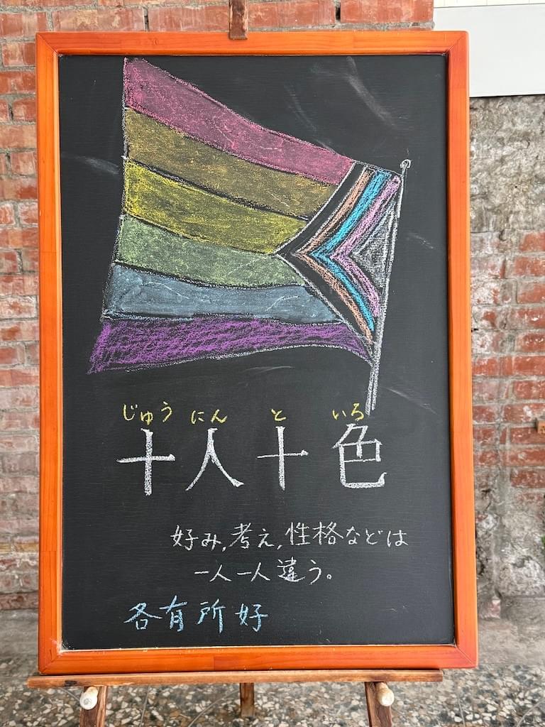 小さい黒板にチョークで絵が描いてあります。日本語のことわざを説明するための黒板です。 上部に大きく、「レインボーフラッグ」のイラストが描いてあります。プログレス・プライドフラッグと呼ばれるタイプ。赤、橙、黄、緑、青、紫の六色の帯に、５色(黒、茶色、水色、ピンク、白)のV字型模様を追加したもの。 はためくフラッグの下に横書きで「十人十色」と書いてあります