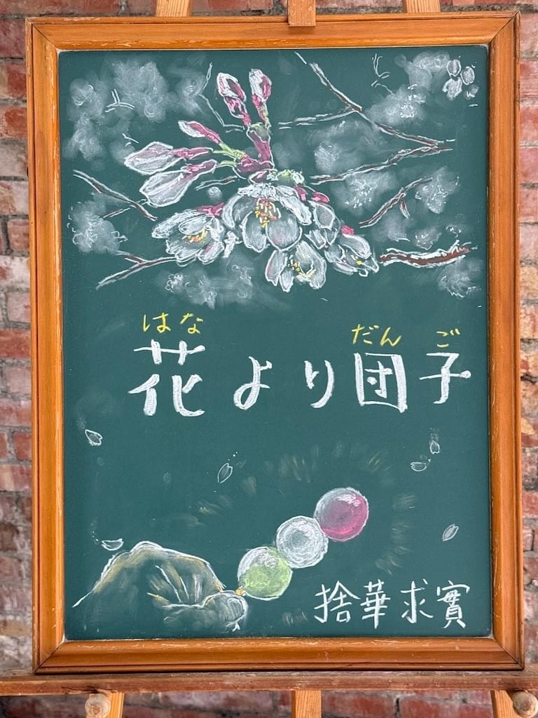 小さい黒板にチョークで絵が描いてあります。日本語のことわざを説明するための黒板です。 縦長の黒板で、上部に桜の花のイラストが描いてあります。 その下に横書き一行で「花より団子」と書いてあります。 下部に左手で、三色団子の串の部分を持っているイラストが描いてあります。団子は下から緑、白、赤です。