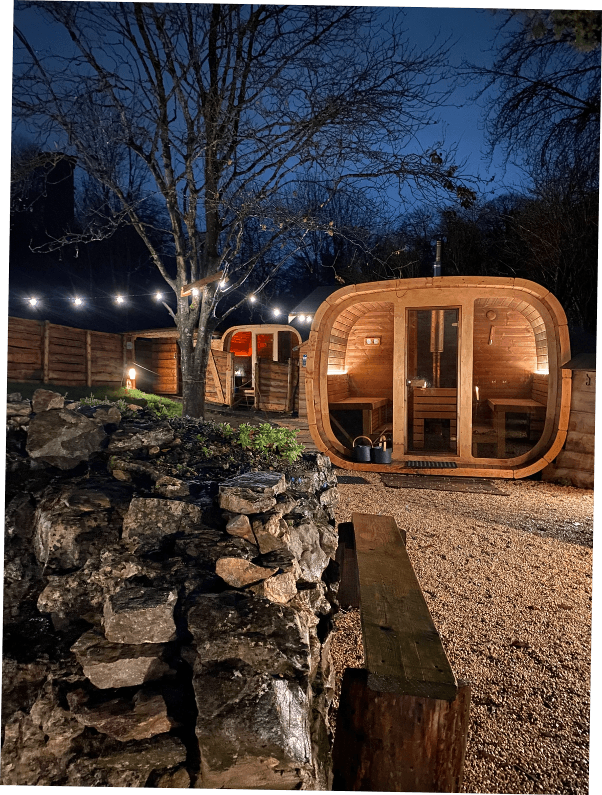 Two saunas glow against a dark winter sky. A bare tree's braches are lit with a string of lightbulbs.
