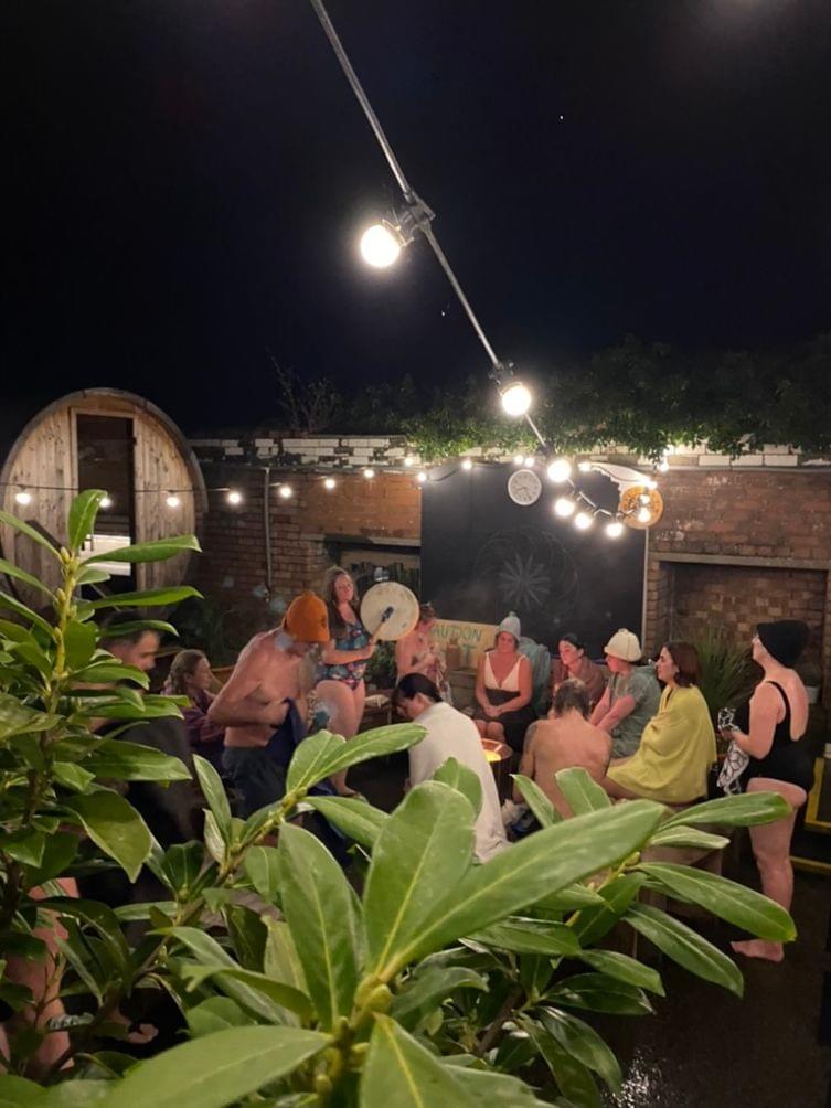 A group in sauna hats, swimmers and towels listen to a drum playing at a sauna ritual.