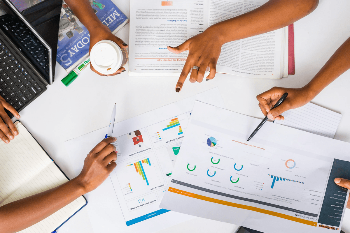 Photo of people pointing at data charts on a table. Photo of people pointing at data charts on a table.