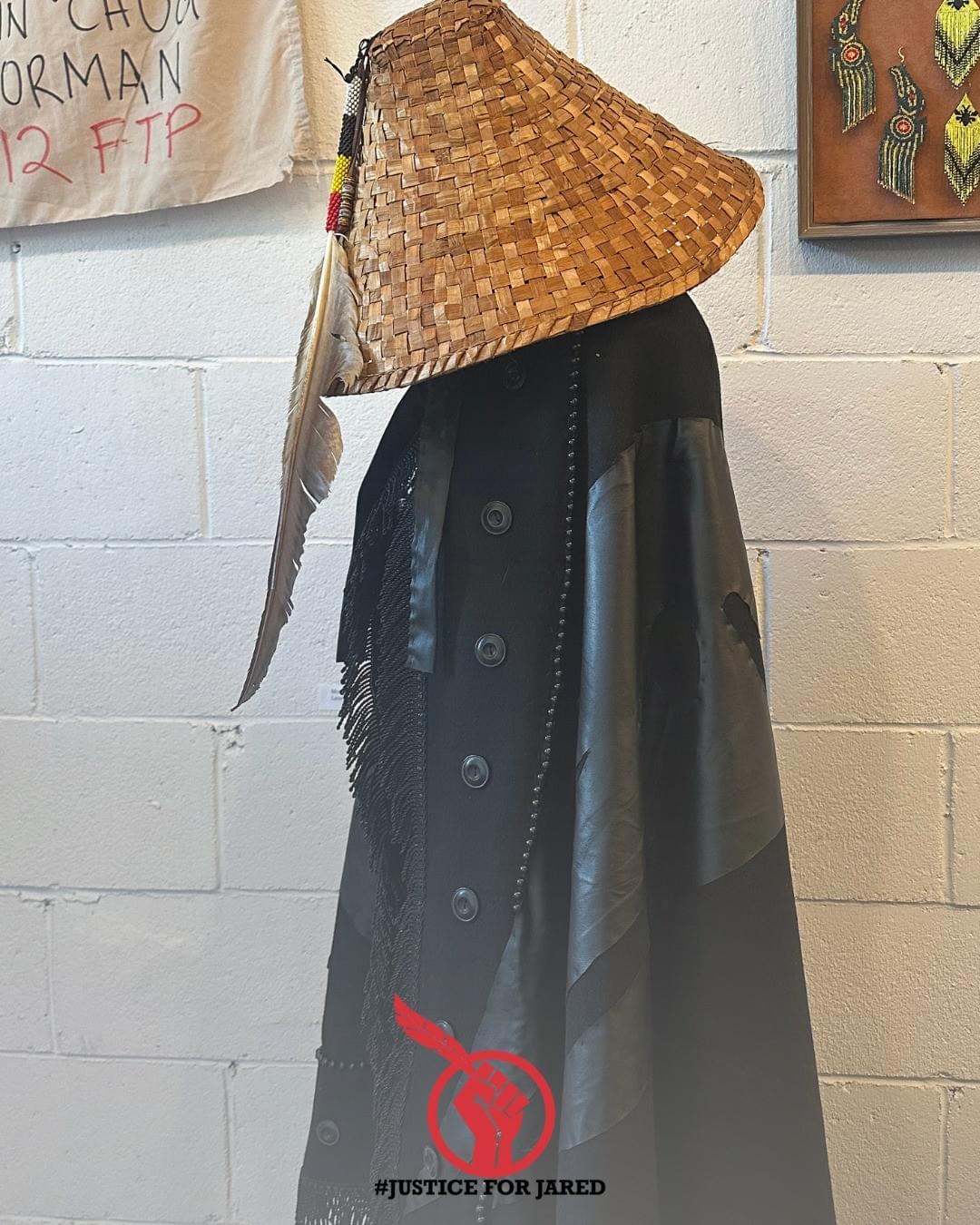 Mourning shawl created by THIRTEEN TWELVE. The mannequin is outfitted with a handwoven cedar har with a large eagle feather adornment. At the bottom of the image is the #JusticeForJared logo, a red fist holding an Eagle feather.
