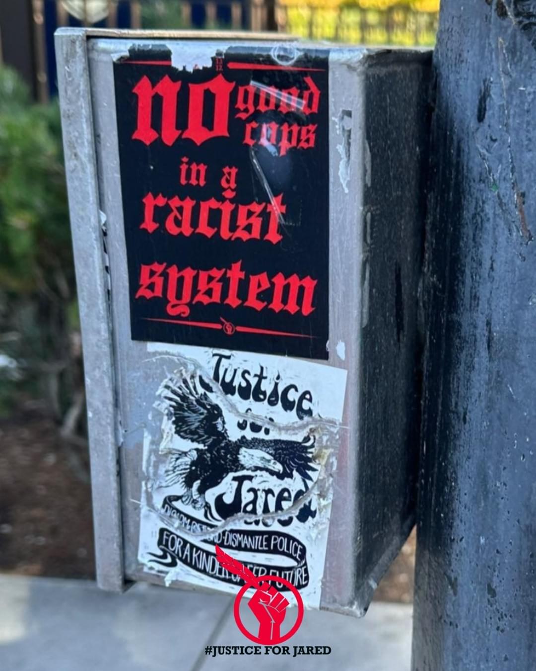 2 Justice for Jared stickers affixed to a metal box. The top sticker reads "no good cops in a racist system. The bottom sticker is a black and white linocut eagle, with the text "Justice for Jared. Disarm, defund, dismantle police for a kinder, safer future." 2 Justice for Jared stickers affixed to a metal box. The top sticker reads "no good cops in a racist system. The bottom sticker is a black and white linocut eagle, with the text "Justice for Jared. Disarm, defund, dismantle police for a kinder, safer future."