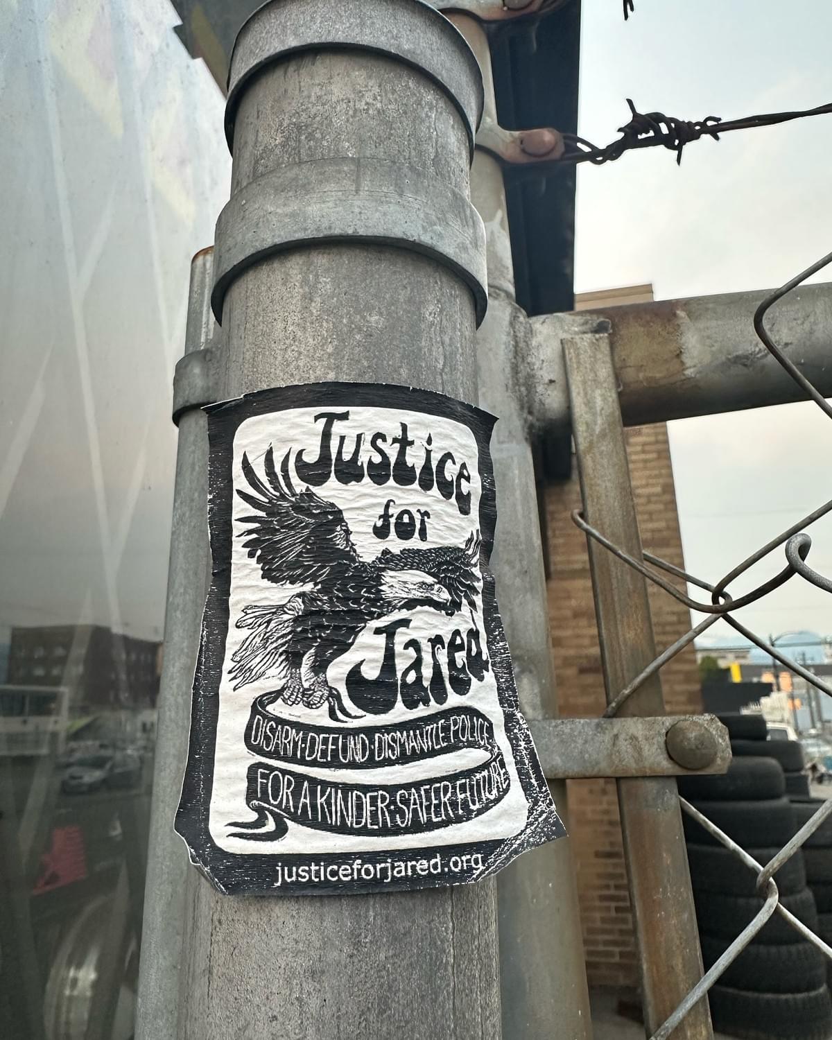 A #JusticeForJared sticker on an east van metal fence. The image is an eagle holding a banner that states “Disarm Defund Dismantle Police. For a Kinder, Safer Future. JusticeForJared.org