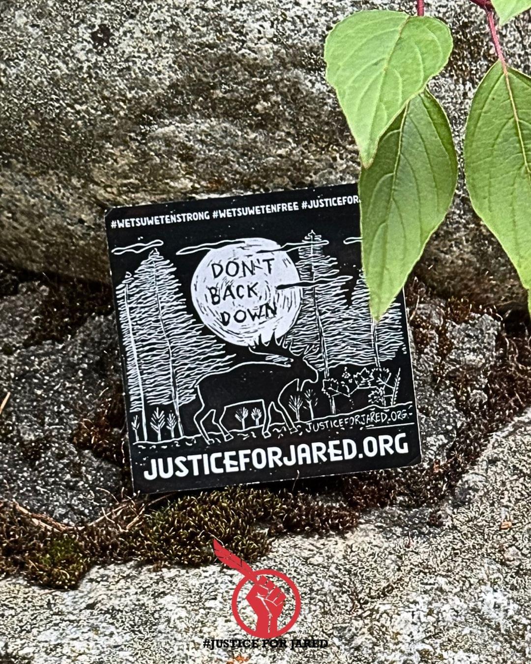 A Justice for Jared sticker photographed at Gott Creek (St'át'imc territory). The black and white sticker features a simple woodcut of a moose under a full moon with the text "WetsuwetenStrong #WetsuwetenFree #JusticeForJared. The sticker is positioned on mossy rocks with a few leaves from a red osier. The Justice for Jared logo is visible at the bottom of the graphic. 