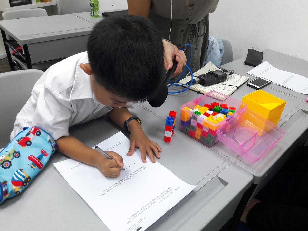 Child writing on worksheet at classroom desk while teacher assists with headphones