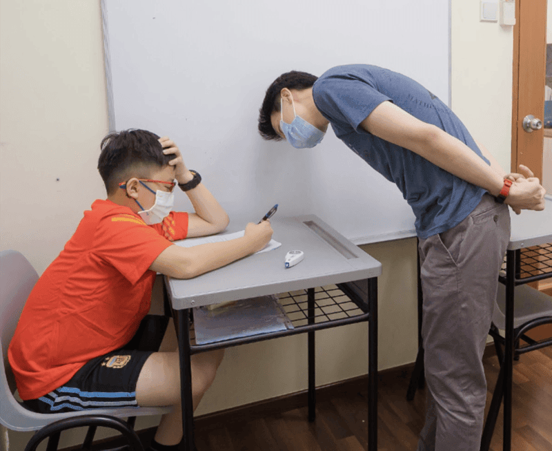 Masked student writing on a worksheet at a classroom desk while an adult instructor leans over to provide individual guidance, with pens, papers, and a whiteboard visible in the study room.