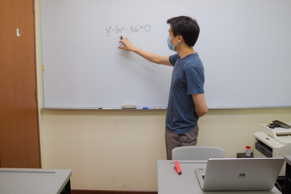 A math tutor wearing a face mask explains a quartic equation on a classroom whiteboard during a one-on-one mathematics tuition lesson.