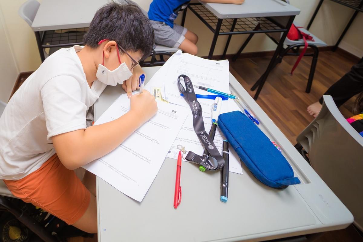 Young student wearing glasses and a face mask completing a mathematics worksheet at a classroom desk, surrounded by pens, stationery, and study materials during a focused learning session.