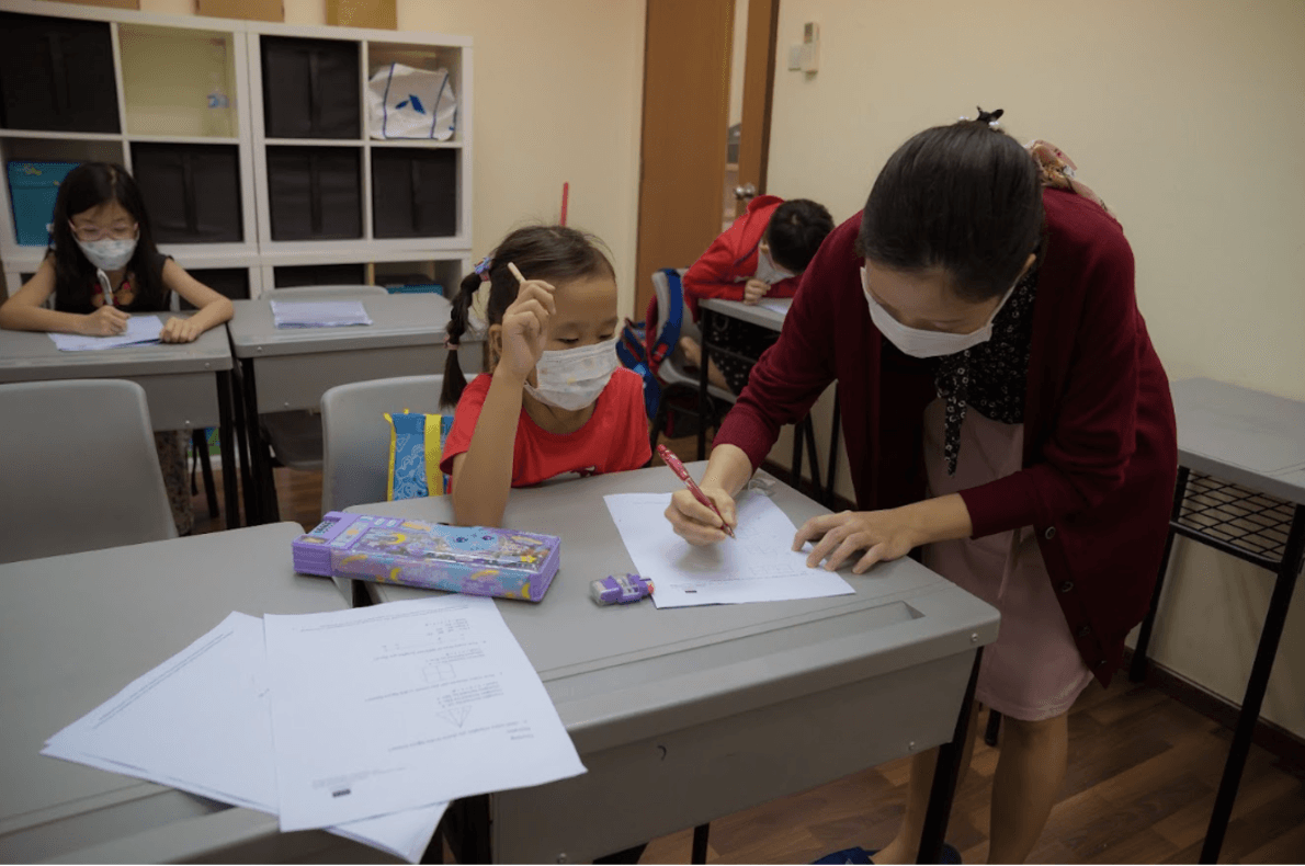 A teacher wearing a face mask assists a young student with a worksheet during a classroom lesson, providing one-on-one guidance at the student's desk.