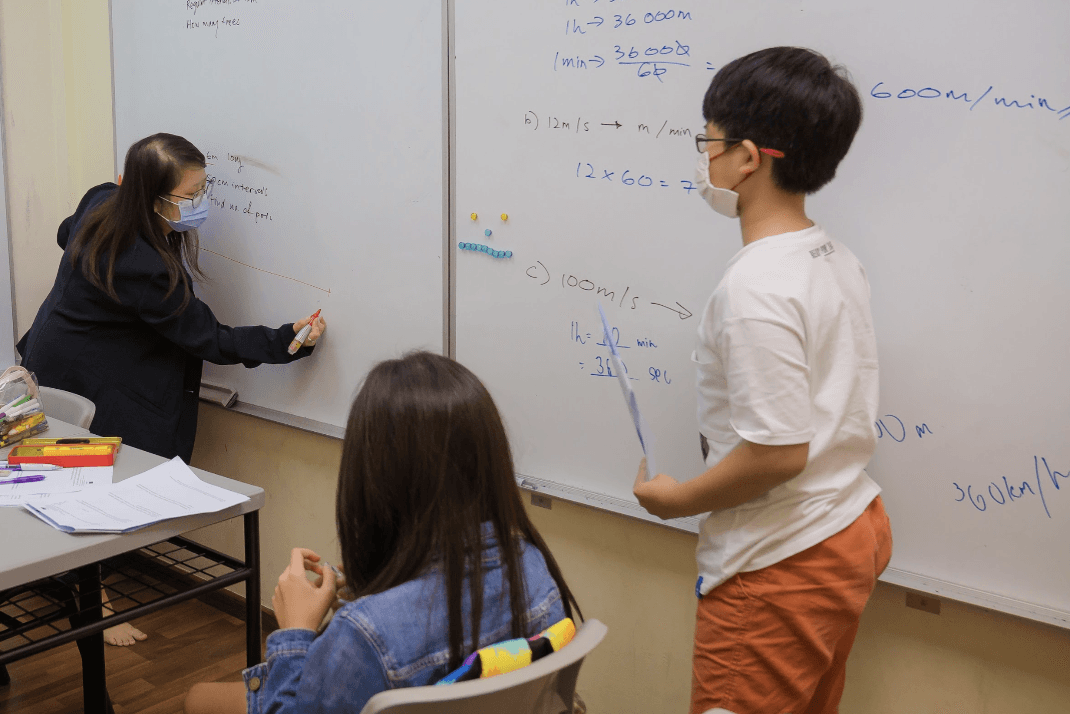 A math tutor helps a student solve measurement and speed-conversion problems on a whiteboard while another student observes in a classroom.