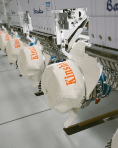 Industrial embroidery machine stitching logos onto cream caps in production line, highlighting custom hat manufacturing.