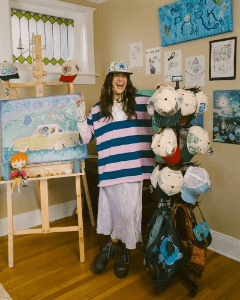 Woman wearing an oversized striped sweater and custom embroidered cap, smiling beside colorful artwork and a display of custom SNAG hats in a creative studio.