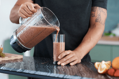 Man pours a berry smoothie from a blender into a glass on a kitchen counter with fresh fruit