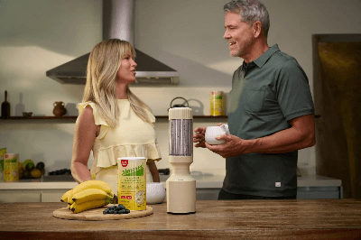 Almased two people talking in modern kitchen with blender, protein container, bananas and blueberries, showcasing balanced nutrition prep