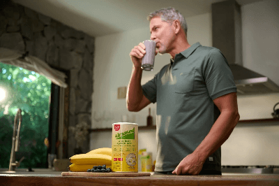 Almased person drinking smoothie in kitchen with protein container, bananas and blueberries, emphasizing balanced nutrition and wellness