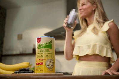 Almased protein container on kitchen counter with bananas and blueberries, person drinking smoothie, emphasizing balanced nutrition wellness