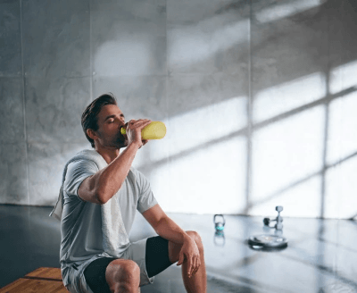 Person rests on a gym bench, sipping from a yellow shaker bottle with towel and gear nearby, emphasizing recovery and hydration.
