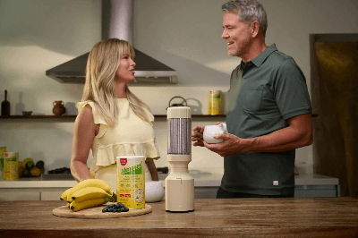 Smiling couple in modern kitchen preparing smoothie with Almased powder, bananas, and blueberries, promoting healthy nutrition.