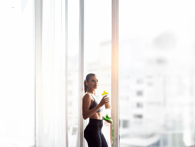 Woman in workout gear holding an Almased shaker and jump rope by a sunlit window.