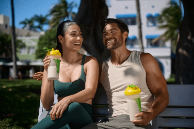 Smiling couple sitting outdoors enjoying protein shakes together, suggesting a healthy lifestyle and shared wellness routine.