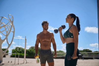 Fit man and woman exercising outdoors under bright blue sky; woman lifts dumbbells while man holds Almased shaker, promoting active lifestyle.