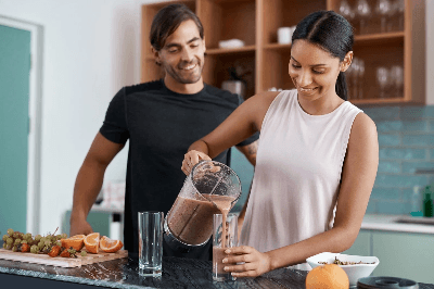 Almased person pouring smoothie from blender into glass with fresh fruit on counter, emphasizing wellness, freshness and healthy living