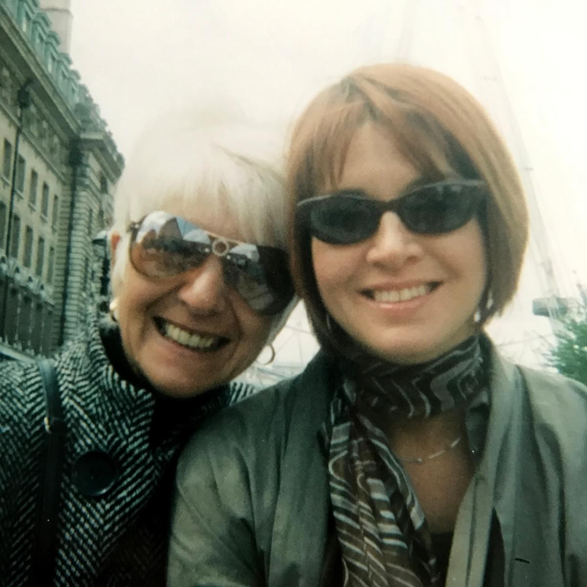 Two women are smiling at the camera. They are mother (LHS) and daughter (RHS). Both are wearing sunglasses, coats and scarves. They are outdoors, in London. Behind the mother is a big, grey building.