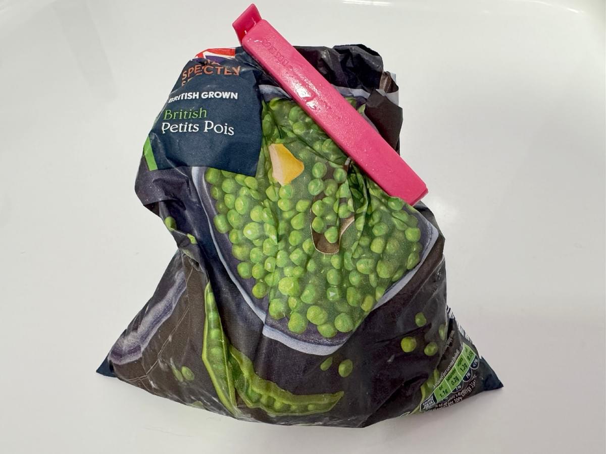 White background. A bag of frozen peas sit crumpled in the centre. A pink, plastic food-clip is attached to the top right hand corner.