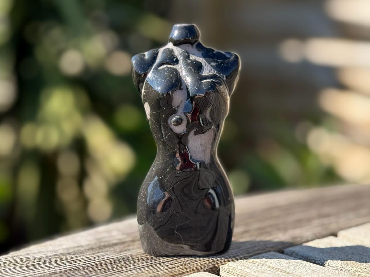 Ceramic torso artwork, black with a bubbled glazing effect. The artwork is placed on a wooden table outdoors. Green shrubbery is blurred in the background