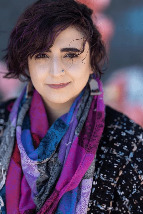 A headshot of Naseem, a light-skinned nonbinary West Asian person with curly brown hair. They're wearing a colorful scarf over a black sweater with threads of blue and cream. They smile with closed lips at the camera.