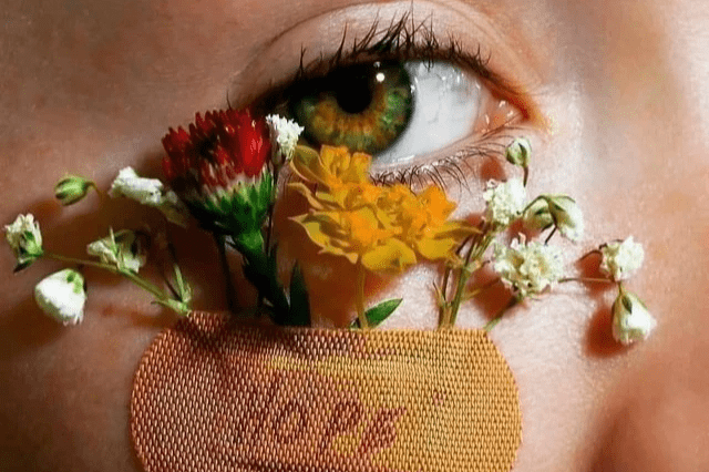 A photo of a hazel eye looking into the camera with a bandaid underneath that reads "HOPE" and white, red and orange flowers sticking out of the bandaid over the eye.