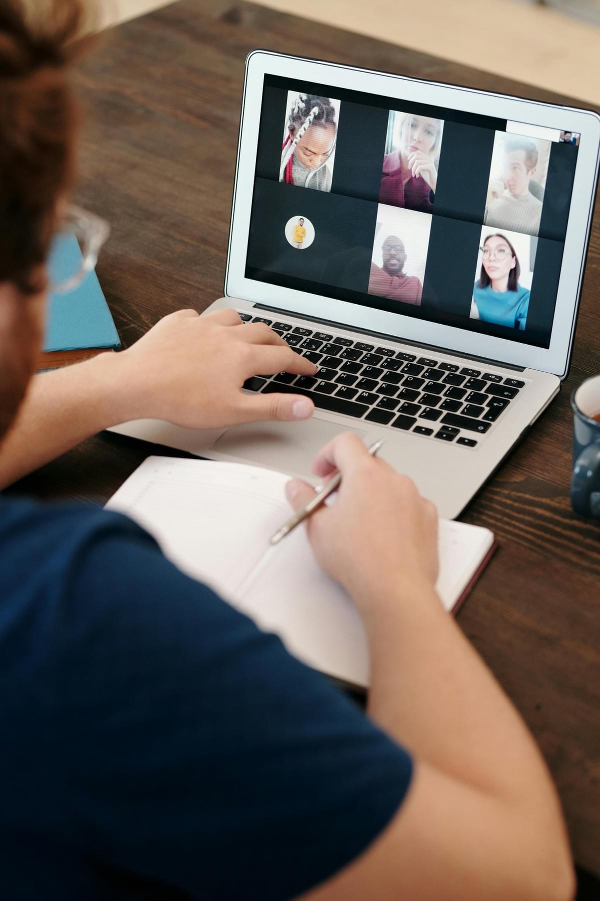 A group chatting among themselves on Zoom shown on a Mac on a desk with a white notebook in front.