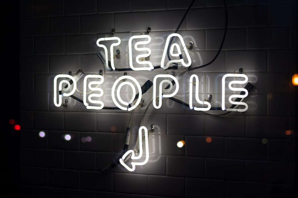 A photo of a white neon sign on a dark wall that reads "TEA PEOPLE" with an arrow pointing left.