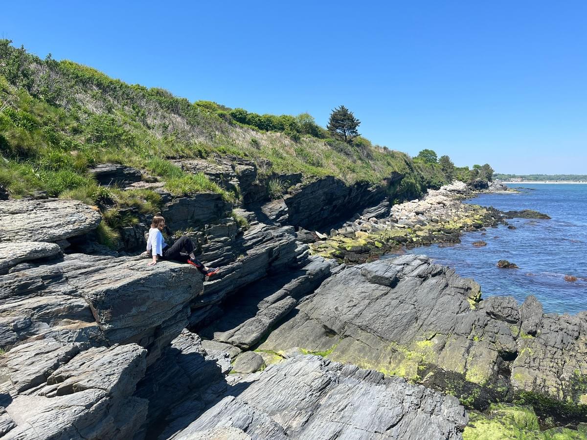 Aleksa sits on a rocky shelf on the Rhode Island shore, gazing at the Atlantic ocean. , bedrocks and the horizon line.