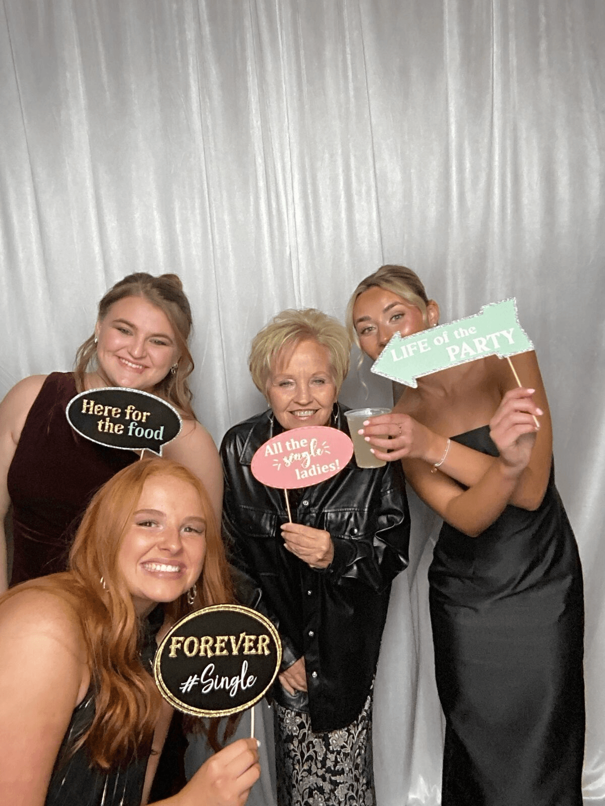 Three wedding guests laugh and hold fun signs at a Tap & Pose booth with a white shimmer curtain