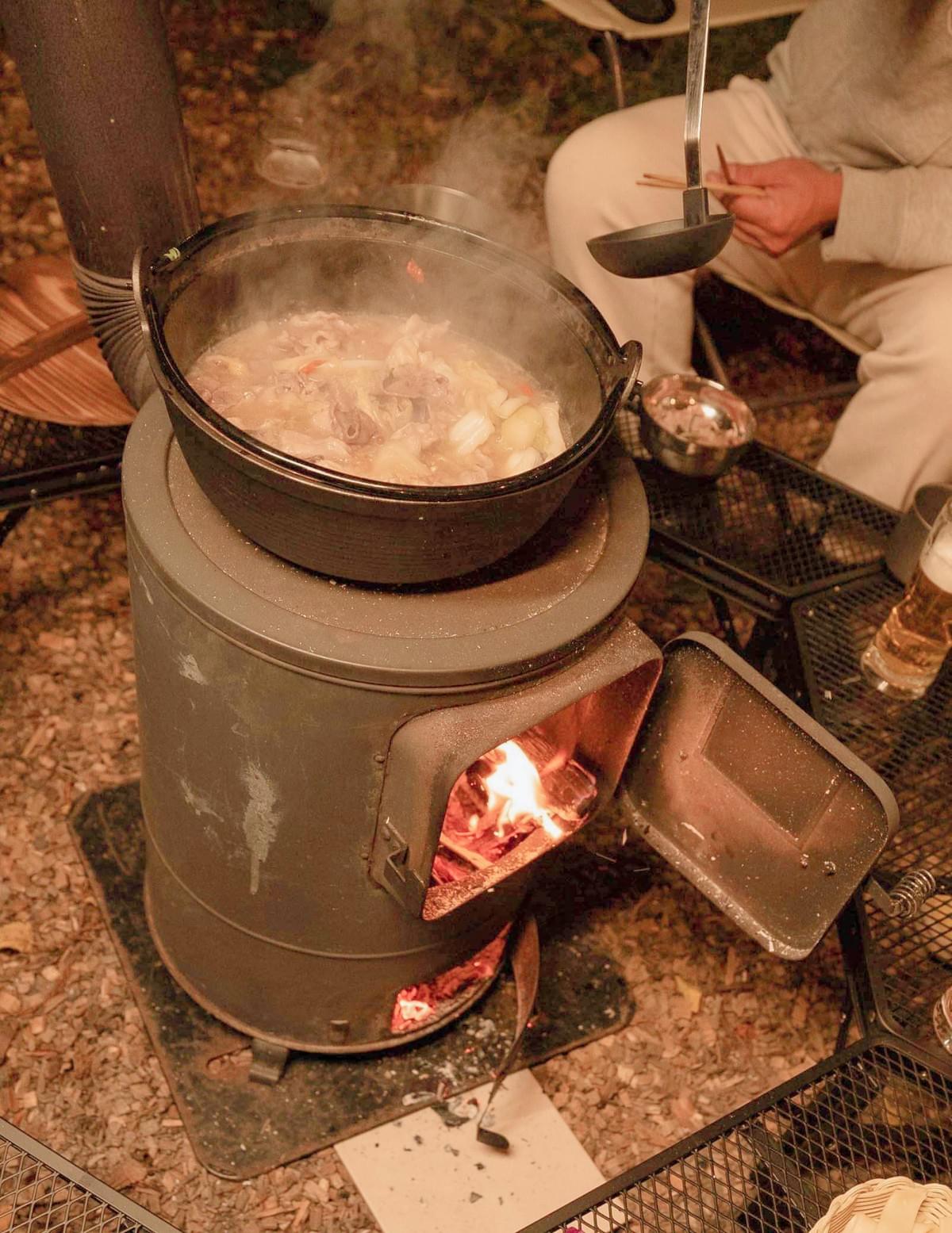 薪ストーブの上に置いた鍋から湯気が立ち、火口に薪の炎が見える