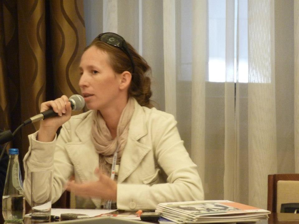 Zdjęcie przedstawiające Iwonę Janicką - uśmiechniętą kobietę w długich włosach spiętych z tyłu ... trzymająca mikrofon w prawej ręce