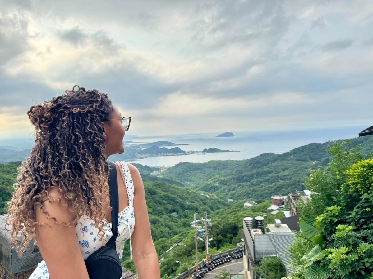 Brit Thompson sitting on ledge at the top of the Juifen Old Street view of the forest and ocean
